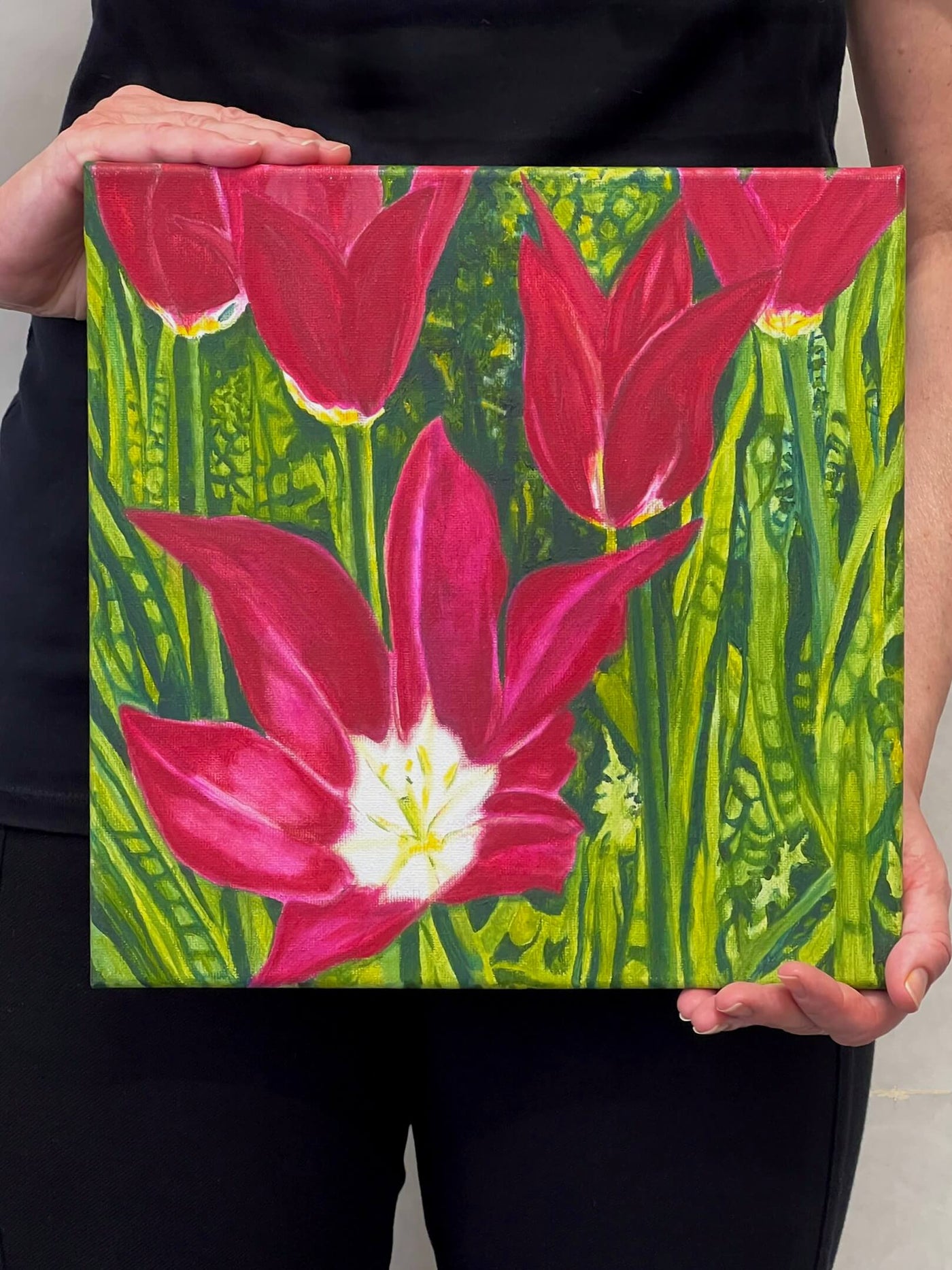 A painting of dark magenta tulips in full bloom, surrounded by a lush, vivid green garden backdrop being held by fine artist Nancy McLennon